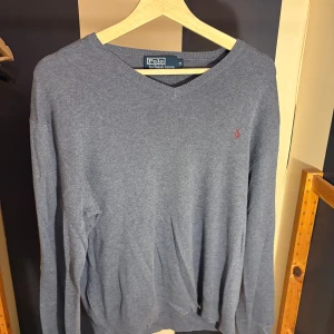 Grå stickad tröja från Polo Ralph Lauren - Snygg grå stickad tröja från Polo Ralph Lauren med v-ringning och diskret broderad logga på bröstet. Långärmad modell i mjukt material som passar perfekt till jeans eller chinos. Klassisk och stilren look.