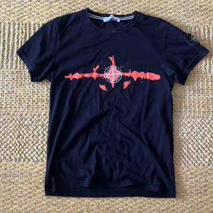 Svart Stone Island t-shirt med tryck - Svart t-shirt från Stone Island med stort rött och vitt tryck på bröstet och tydlig logga på ärmen. Baksidan har STONE ISLAND i stora vita bokstäver. Klassisk rund hals och korta ärmar, tillverkad i mjuk bomull. Perfekt för dig som gillar streetwear och vill sticka ut.