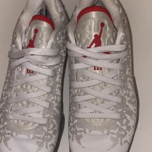 Vita Air Jordan sneakers med röd logga - Unika vita Air Jordan sneakers med grått mönster och röd Jumpman-logga på plösen. Skorna har vita snören, gråmelerad sula och svart blixtdetalj på hälen. Ovandelen är i syntetmaterial med cool struktur och detaljer som sticker ut.