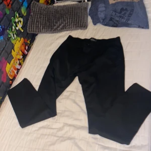 Svarta kostymbyxor Tailored Originals - Snygga svarta kostymbyxor från Tailored Originals i klassisk rak modell. Byxorna har en stilren look och passar perfekt till en dressad outfit. Materialet känns mjukt och följsamt, vilket gör dem bekväma att bära hela dagen.