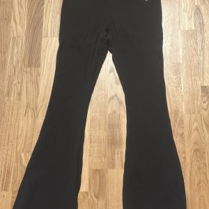 Svarta ribbade bootcut-byxor Nike M - Svarta ribbade byxor från Nike i bootcut-modell. Byxorna har elastisk midja och är tillverkade i ett mjukt, stretchigt material. Snyggt broderad Nike-logga framtill. Perfekta för en chill och sportig look.