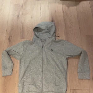 Grå hoodie från Polo Ralph Lauren - Snygg grå hoodie från Polo Ralph Lauren med dragkedja och klassisk broderad logga på bröstet. Tröjan har huva med snörning, två fickor framtill och är tillverkad i mjuk bomull. Perfekt för en chill och stilren look. Köpt i canada