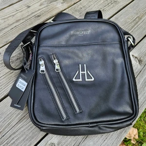 Svart läder axelväska från Horspist Paris - Snygg svart läder axelväska från Horspist Paris i skinn med två sneda dragkedjor framtill och silverfärgad logga. Väskan har justerbar axelrem och stilren, fyrkantig form. Perfekt för att bära dina essentials med stil.