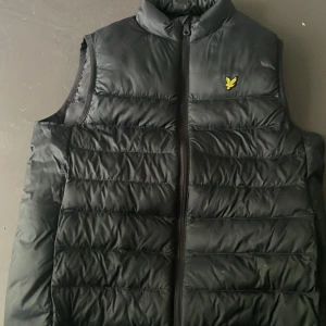 Svart väst från Lyle & Scott 12 - 13 år - Svart väst från Lyle & Scott med klassisk gul logga på bröstet. Västen är quiltad med horisontella sömmar och har hög krage samt dragkedja framtill. Perfekt att slänga över en hoodie för en chill look.