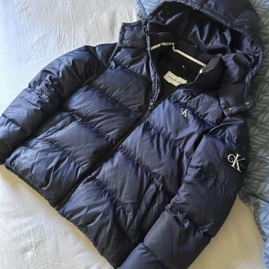 Svart pufferjacka från Calvin Klein - Säljer en mörkblå pufferjacka från Calvin Klein Jeans med huva och dragkedja. Nypris ca (2500kr). Jackan har en broderad CK-logga på ärmen och en patch på bröstet. Fodrad insida och två fickor framtill. Perfekt för kalla dagar och riktigt snygg jacka!