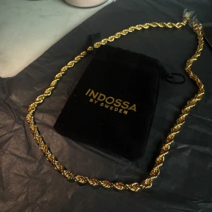 Cordell Halsband 6 mm 60 cm 18k GuldPläterad  - Snyggt guldfärgat halsband från Indossa by Sweden med tvinnad kedja och glittrig låsdetalj. Perfekt accessoar för att lyfta din outfit med en lyxig touch. Kommer med svart sammetsförpackning med guldig logga.            Aldrig använt, plasten sitter kvar runt kistlåset!  I rostfritt stål så den ändrar ej färg och går att ha på sig när man duschar och badar :) halsbandet är 6 mm bred och 60 cm lång. 