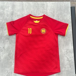 Röd Spanien fotbollströja H&M nr 10 - Snygg röd fotbollströja från H&M Sport med gula detaljer och tryck. Nummer 10 på både fram- och baksida samt Spanien-emblem på bröstet. Kortärmad modell med gul kant vid ärmslut och hals. Tillverkad i lätt och ventilerande polyester, perfekt för match eller träning.