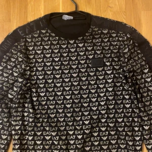 Svart EA7 långärmad tröja med logga - Säljer en svart långärmad tröja från EA7 Emporio Armani med all-over logomönster i vitt. Tröjan har rund halsringning och ribbade muddar. Perfekt för dig som gillar streetwear och vill sticka ut med en snygg designertröja.