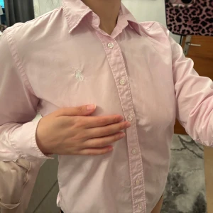  skjorta  Ralph Lauren - Säljer en klassisk ljusrosa skjorta från Ralph Lauren Sport med vit broderad logga på bröstet. Skjortan har lång ärm, knappar hela vägen och en snygg krage. Tillverkad i bomull och har en figurnära passform som ger en clean och stilren look.