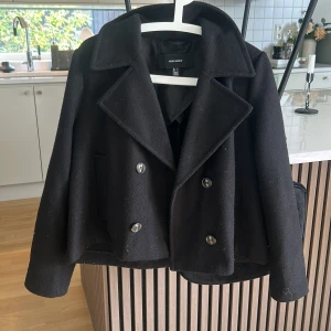 Svart kappa från Vero Moda - Stilren svart kappa från Vero Moda med dubbelknäppning och bred krage. Klassisk look som funkar till många outfits. Original pris 600 kr 