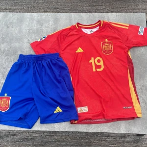 Spanien Lamine Yamal #19 Adidas matchtröja - Säljer en officiell Spanien fotbollströja från Adidas med Lamine Yamal och nummer 19 på ryggen. Tröjan är röd med gula detaljer, korta ärmar och broderat klubbmärke. Materialet är lätt och andas, perfekt för match eller träning. Ingår blå shorts med gul logga och klubbmärke.