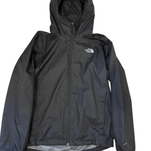 Svart vindjacka från The North Face - Svart vindjacka från The North Face med huva och dragkedja framtill. Jackan har meshfoder på insidan och klassisk logga på bröstet. Perfekt för blåsiga dagar och enkel att slänga på sig när vädret svänger. Storlek L