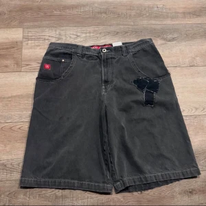 Svarta loose shorts från JNCO Jeans - Riktigt feta svarta shorts från JNCO Jeans med loose fit och breda ben. De har coola detaljer som patchwork på framsidan och en stor broderad bulldogg på bakfickan. Klassisk femficksmodell i jeansmaterial, perfekta för dig som gillar streetwear och vill sticka ut.