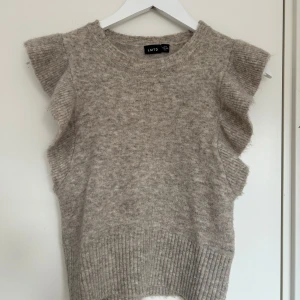 Beige stickad topp från LMTD - Säljer en mysig beige stickad topp från LMTD. Den har volangdetaljer vid ärmarna och en ribbad nederkant. Perfekt för en stilren och bekväm look. Passar bra till både jeans och kjol.