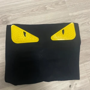 Svart kuvertväska med gula detaljer - Fendi T-shirt Cool svart kuvertväska med två stora gula ögon framtill som ger en edgy look. Väskan är rektangulär och har en slät yta med detaljer i plast. Perfekt för dig som vill sticka ut med en unik accessoar.