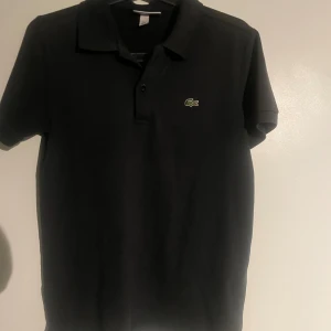 Svart Lacoste slim fit piké M - Svart pikétröja från Lacoste i slim fit-modell med klassisk krage och två knappar framtill. Ikoniska gröna krokodilloggan på bröstet. Tillverkad i mjuk bomull som är skön mot huden. Perfekt för en clean och sportig look.  Använd fåtal gånger 9/10 skick