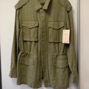Olivgrön parkajacka från Gina Tricot - Snygg olivgrön parkajacka från Gina Tricot i linneblandning. Jackan har fyra stora fickor med lock, axelklaffar och dragsko i midjan för en cool passform. Perfekt för dig som gillar utility-vibe och vill ha en jacka med mycket detaljer. Helt ny.