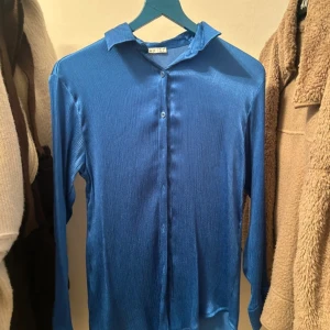 Blå glansig skjorta med knappar - Snygg blå skjorta med glansigt tyg och diskret ribbad struktur. Skjortan har klassisk krage, långa ärmar och knäppning framtill. 