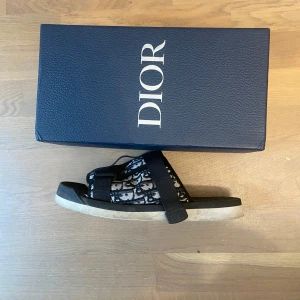 Svarta sandaler från Dior - Snygga sandaler från Dior med svart och beige mönstrad textil och breda svarta remmar. Sulan är platt och svart med vit undersida, och ovandelen har tydligt Dior-logomönster. Perfekta för sommaren och ger en lyxig touch till din outfit. Låda till sandalerna kommer med