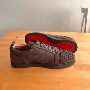 Christian Louboutin sneakers med nitar - Snygga mörkgrå sneakers från Christian Louboutin i mocka med ikoniska röda sulor. Skorna har rund tå, snörning och coola svarta nitar framtill som ger en edgy vibe. Perfekta för dig som vill sticka ut med lyxiga detaljer och streetstyle-känsla.