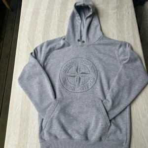 Grå hoodie från Stone Island - Snygg grå hoodie från Stone Island med stort broderat logotyp på bröstet. Klassisk känguruficka framtill, ribbade muddar och huva med metallöljetter. Perfekt för dig som gillar streetwear och vill ha en stilren look. Lite små noppig.