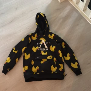 Skidjacka Slade Couture  med gummiankor - Svart jacka  från Slade Couture med stora gula gummiankor över hela. Jackan har huva, lång ärm och en praktisk ficka framtill med knappdetalj. Perfekt för dig som gillar unika och lekfulla plagg med coolt tryck. Ficka för liftkort på ärmen.