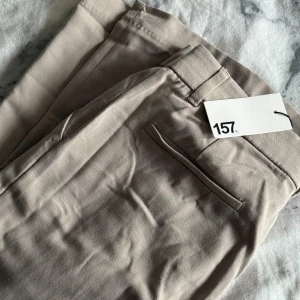Beige kostymbyxor från 157 - Snygga beige kostymbyxor från 157 med klassisk rak passform och diskreta fickor bak. Byxorna har hällor för bälte och är gjorda i ett mjukt bomullsmaterial som känns skönt mot huden. 