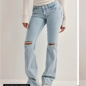 Ljusblå bootcut jeans med slitningar - Snygga ljusblå jeans med bootcut-modell och slitna detaljer vid knäna. Jeansen har klassiska fem fickor och coola broderade bakfickor med zigzag-mönster. Perfekta för en avslappnad och trendig look. använd fåtal gånger, har även sprättat upp byxorna då de va lite korta för mig men enkelt att lägga upp igen om man vill