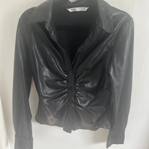 Svart skinnskjorta från Zara, M - Snygg svart skjorta från Zara i storlek M, tillverkad i skinnimitation. Skjortan har lång ärm, klassisk krage och knappar framtill. Framsidan är snyggt rynkad för extra edge. Perfekt för dig som gillar en cool och trendig stil. Knapparna är lite lösa men den går att använda samt det går att laga. Lite trådar i vissa sömmar. 