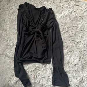 Svart omlottopp från Gina Tricot - Svart omlottopp från Gina Tricot i mjuk polyester. Toppen har långa ärmar och en snygg knytning framtill som ger en cool och trendig look. Perfekt för dig som gillar stilrena plagg. Passar dig som vill ha en slim passform.