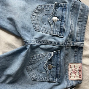 True Religion Joey ljusblå flare jeans - Säljer ett par ikoniska True Religion Joey jeans i ljusblå tvätt med snygga kontrastsömmar och stora fickor bak. Modellen är low rise med flare och har slitna detaljer vid benslut. Perfekta för dig som gillar statement-jeans med vintagevibe. Lite slitna nere vid fötterna då de är rätt så långa på mig annars inga defekter 🥰💞