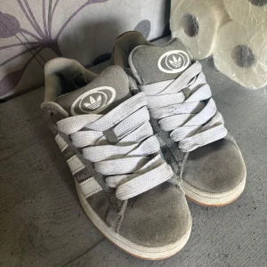 Grå Adidas sneakers i mocka - Snygga grå sneakers från Adidas med klassiska vita ränder på sidorna och stora loggor på plös och häl. Skorna har breda vita snören och ovandel i mjuk mocka. Perfekta för dig som gillar en clean och sportig stil.