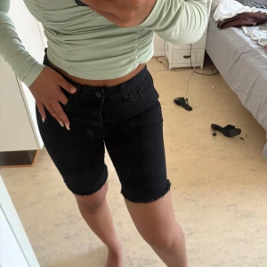 Svarta jeansshorts med rå kant - Säljer ett par svarta jeansshorts med midwaist passform och rå, fransad kant vid benslut. Modellen är normal i passningen och har klassiska fickor fram och bak samt knapp och dragkedja. Perfekta för dig som gillar en enkel och clean look.