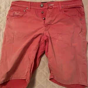 Rosa shorts från Jacob Cohen - Snygga röda shorts från Jacob Cohen med klassisk femficksdesign och vita kontrastsömmar. Modellen är lite längre och har knäppning med tre knappar framtill. Perfekta för sommardagar när du vill sticka ut lite extra.