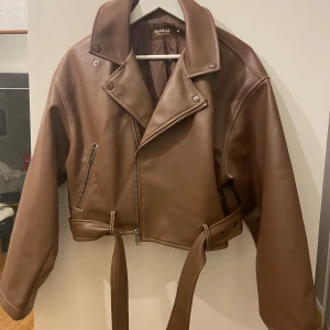 Brun skinnjacka från Bianca x Nelly - Säljer en brun bikerinspirerad skinnjacka från Bianca x Nelly. Jackan är croppad med bred krage, dragkedja framtill, bälte i midjan och dragkedjor vid ärmsluten. Perfekt för dig som vill ha en edgy och trendig look.