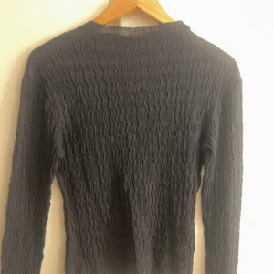 Svart långärmad ribbad tröja - Svart långärmad tröja med coolt ribbat och vågigt strukturmönster. Sitter tajt mot kroppen och har en enkel, stilren design. Båtringning i halsen 