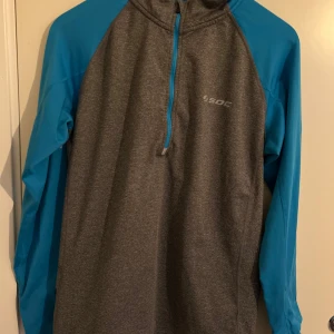 Grå och blå träningshoodie från SOC - Sportig hoodie från SOC i grått med blå ärmar och detaljer. Tröjan har lång ärm, half zip framtill och är tillverkad i ett syntetmaterial som andas. Perfekt för träning eller löprundan när det är lite kyligare ute.