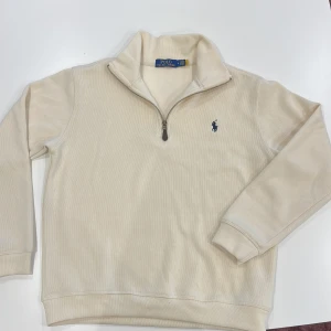Beige half zip tröja Polo Ralph Lauren - Snygg beige långärmad tröja från Polo Ralph Lauren med half zip och klassisk blå logga broderad på bröstet. Tröjan har hög krage och är gjord i mjuk bomull, perfekt för lager-på-lager. Enkel och stilren design som funkar till allt. Passar S använd en gång
