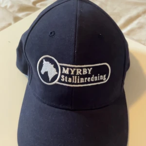 Mörkblå keps Myrby Stallinredning - Snygg mörkblå keps med vit broderad häst och texten 'MYRBY Stallinredning' framtill. Klassisk böjd skärm och justerbar baktill. Perfekt för dig som gillar hästar och vill ha en stilren look.