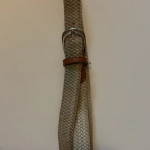 Beige flätat bälte med skinn-detaljer - Snyggt beige flätat bälte med bruna skinn-detaljer och silverspänne. Perfekt accessoar för att lyfta din outfit med en stilren och avslappnad känsla. Bältet har en klassisk design som passar till många olika looks.