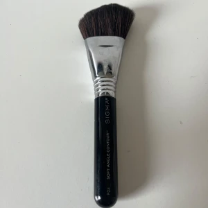 Sigma Soft Angle Contour F23 borste - En sminkborste från Sigma, modell Soft Angle Contour F23. Borsten har ett svart handtag, silverfärgat fäste och mjuka, täta borst i brunt. Perfekt för contouring och applicering av bronzer eller rouge med precision.
