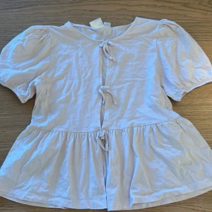 Vit blus med knyt från Gina Tricot storlek  - Supersöt vit blus från Gina Tricot i storlek 170. Blusen har puffiga korta ärmar, volang nedtill och tre knyt framtill som ger en gullig vibe. Tillverkad i mjuk bomull, perfekt för dig som gillar romantisk och chill stil.