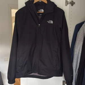 Svart vindjacka från The North Face - Svart vindjacka från The North Face med huva och dragkedja framtill. Jackan har meshfoder på insidan, elastiska muddar och två fickor med dragkedja. Perfekt för blåsiga dagar och enkel att matcha med allt.
