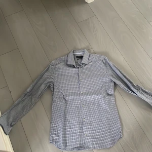 Rutig blåvit skjorta från Riley - Snygg rutig skjorta från Riley i blått och vitt. Slim fit-modell med klassisk krage och långa ärmar. Skjortan har knappar framtill och är tillverkad i ett mjukt bomullstyg. Perfekt för dig som gillar stilrena och fräscha plagg. Perfekt skick