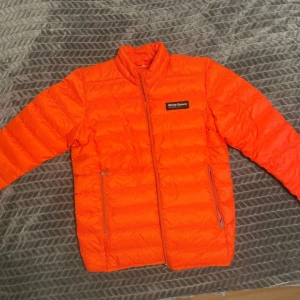 Orange pufferjacka från White Space - Säljer en riktigt snygg orange pufferjacka från White Space, modell Tate Jacket Coral. Jackan har dragkedja framtill, två sidofickor med dragkedja och en clean look med logga på bröstet. Perfekt för dig som vill sticka ut i höst och vinter!