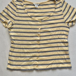Randig ribbad topp från Monki - Superfin ribbad topp från Monki i ljusgul med marinblå och vita ränder. Modellen har korta ärmar, v-ringning och knappar framtill. Perfekt för dig som gillar en chill och trendig stil med lite retrovibbar.
