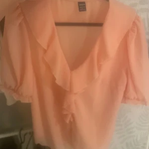 Persikofärgad blus med volang - Söt persikofärgad blus från Shein med v-ringning och volangdetaljer längs halsringningen. Blusen har korta puffärmar med resår vid ärmslut och är tillverkad i ett lätt, luftigt material. Perfekt för dig som gillar romantiska och feminina plagg.
