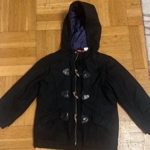 Svart rock med huva från Okaou - Svart rock från Okaou med huva och snygga träknappar framtill. Jackan har två fickor med lock och är helfodrad med mörkblått innerfoder. Klassisk design med lång ärm och stilrena detaljer som passar dig som gillar enkel men cool look.