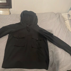 Svart vindjacka från Stone Island - Svart vindjacka från Stone Island med huva, två stora fickor framtill och klassisk patch på vänster ärm. Jackan har dragsko vid halsen och är tillverkad i ett slätt syntetmaterial. Perfekt för dig som gillar streetwear och vill ha en clean look.                 Såklart är det inte ett fast pris😁