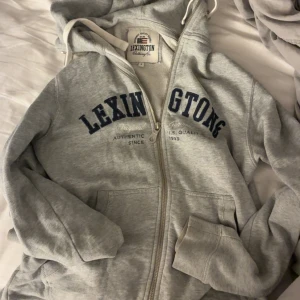 Grå hoodie från Lexington med dragkedja - Snygg grå hoodie från Lexington med marinblå text på bröstet. Tröjan har huva med snörning, dragkedja framtill och två fickor. Perfekt för chill dagar eller när du vill ha en sportig look. Nypris runt 1500kr och modell säljs inte längre 